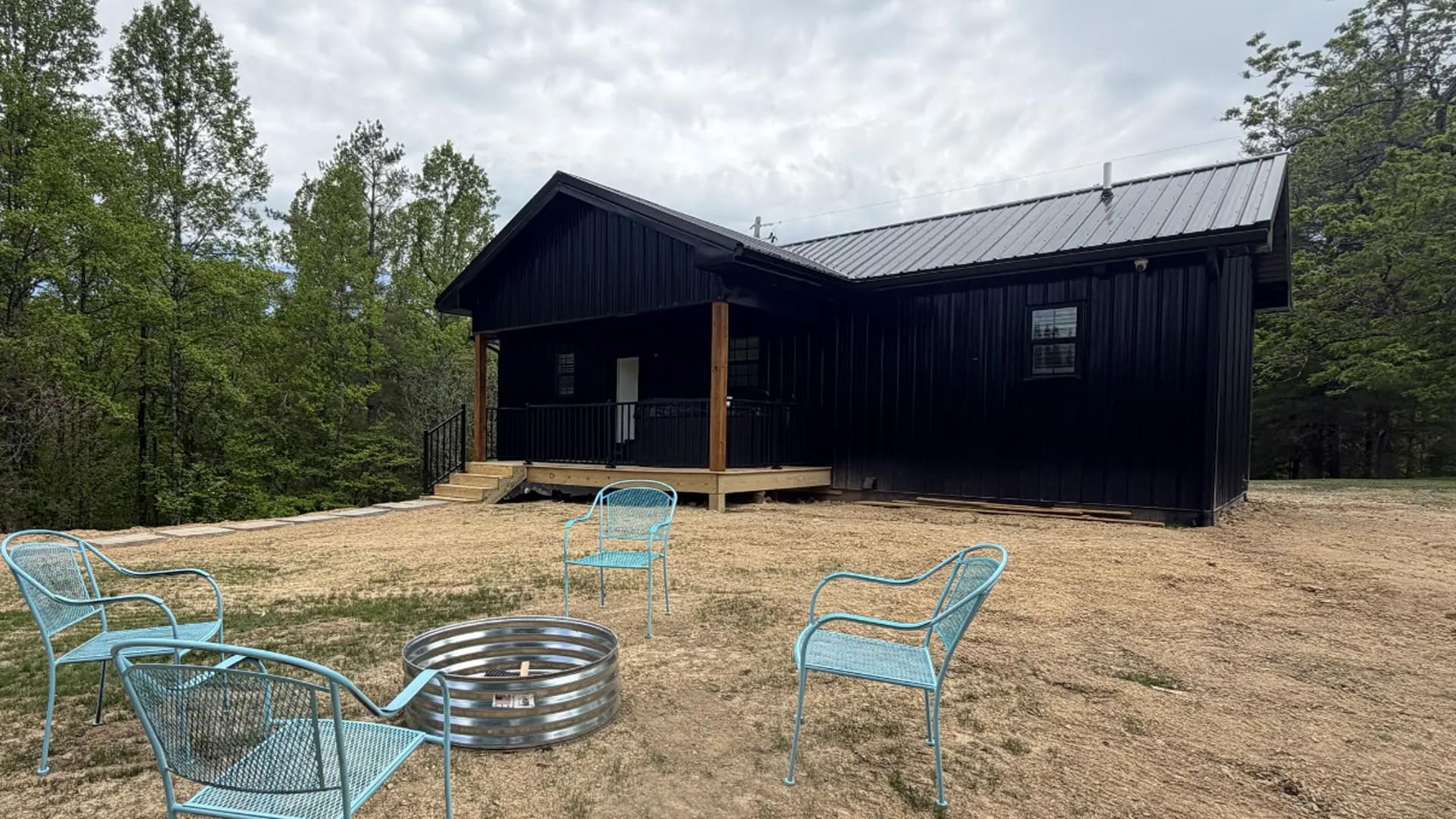 Black Cabin with Metal Roof and Wooden Porch in a Clearing, Blue Metal Chairs Arranged Around a Fire Ring Outdoors.