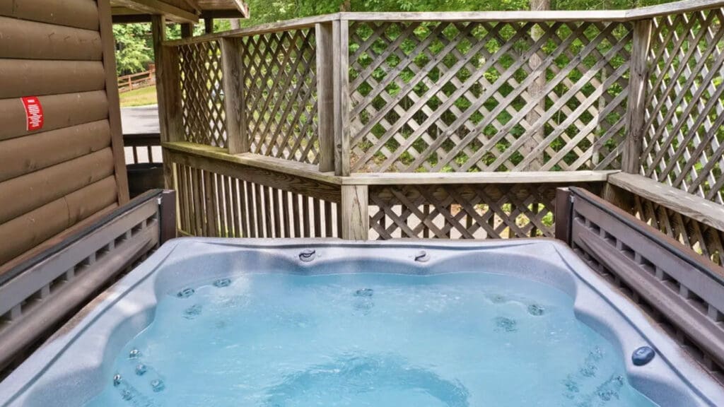 Outdoor Hot Tub on a Wooden Deck with Lattice Privacy Fencing and Green Trees in the Background, Filled with Blue Water and Jets Visible on the Surface.