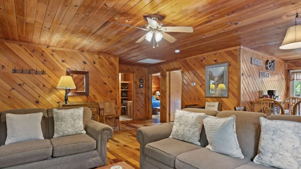 Cozy Cabin Living Room with Wood-paneled Walls, Beige Sofas, and a Ceiling Fan Overhead.